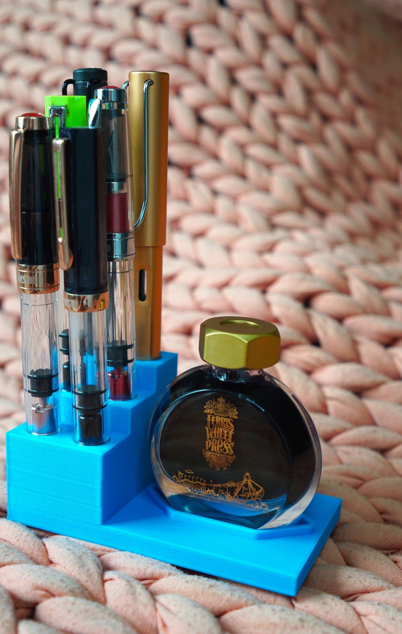 Puede incluir: Un portaplumas azul con cuatro plumas y una botella de tinta Ferris Wheel Press. La botella de tinta es de vidrio transparente con una tapa dorada y una etiqueta negra con letras doradas. La etiqueta dice "Ferris Wheel Press".