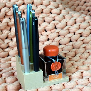 Puede incluir: Un portaplumas de madera con ocho plumas y una botella de tinta. Las plumas son de varios colores, incluyendo plata, marr&oacute;n, verde y azul. La botella de tinta es de vidrio transparente con una tapa roja y una etiqueta que dice "Caran d'Ache #798  Les Ecrivains  Emerald Green".