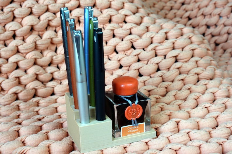 Puede incluir: Un portaplumas de madera con ocho plumas y una botella de tinta. Las plumas son de varios colores, incluyendo plata, marr&oacute;n, verde y azul. La botella de tinta es de vidrio transparente con una tapa roja y una etiqueta que dice "Caran d'Ache #798  Les Ecrivains  Emerald Green".