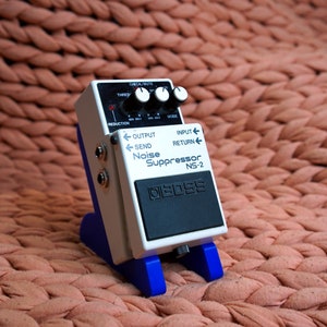 May include: A white Boss NS-2 Noise Suppressor guitar pedal with black knobs and a black foot switch. The pedal is on a blue stand.