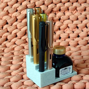 Puede incluir: Un portaplumas blanco con cinco plumas y un frasco de tinta. Las plumas son plateadas, doradas, negras y de oro rosa. El frasco de tinta es azul con una tapa dorada y una etiqueta que dice "J. Herbin 1670 Perle Noire".