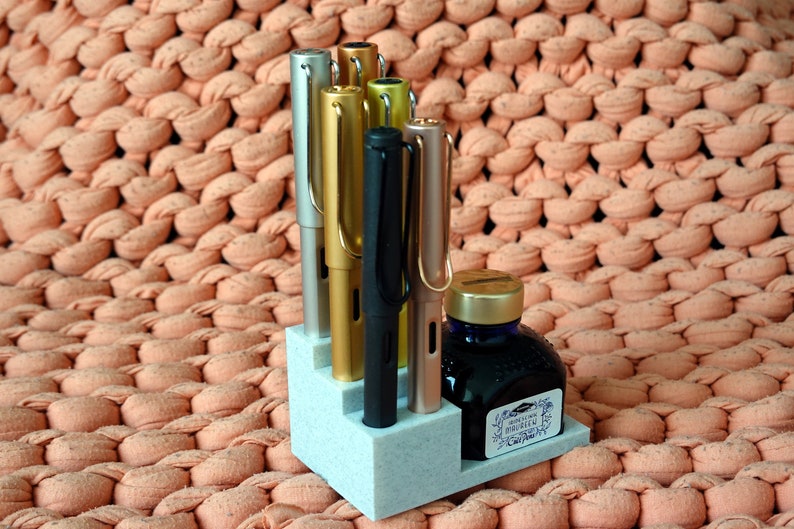 Puede incluir: Un portaplumas blanco con cinco plumas y un frasco de tinta. Las plumas son plateadas, doradas, negras y de oro rosa. El frasco de tinta es azul con una tapa dorada y una etiqueta que dice "J. Herbin 1670 Perle Noire".