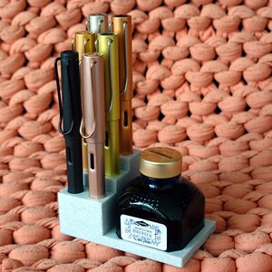 Puede incluir: Un portaplumas gris con cinco plumas, cuatro de ellas son de color dorado met&aacute;lico, oro rosa y plata, y una es negra. Un frasco de tinta azul con tapa dorada est&aacute; delante del portaplumas. El portaplumas y el frasco est&aacute;n sobre una superficie de punto de color melocot&oacute;n.