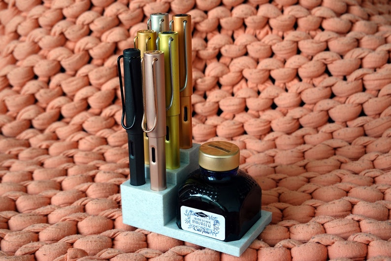 Puede incluir: Un portaplumas gris con cinco plumas, cuatro de ellas son de color dorado met&aacute;lico, oro rosa y plata, y una es negra. Un frasco de tinta azul con tapa dorada est&aacute; delante del portaplumas. El portaplumas y el frasco est&aacute;n sobre una superficie de punto de color melocot&oacute;n.