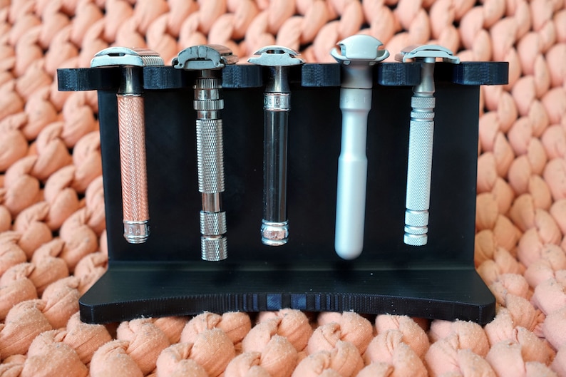 May include: A black plastic stand with five silver and rose gold safety razors. The stand has a textured surface and is designed to hold the razors upright.