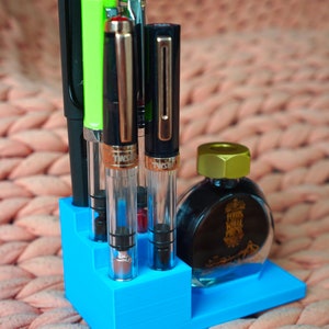 Puede incluir: Un portaplumas de pl&aacute;stico azul con cinco plumas y un frasco de tinta. Las plumas son negras, verdes, plateadas y doradas. El frasco de tinta es transparente con una tapa dorada y dice "Herbs Hill Press" en la etiqueta.