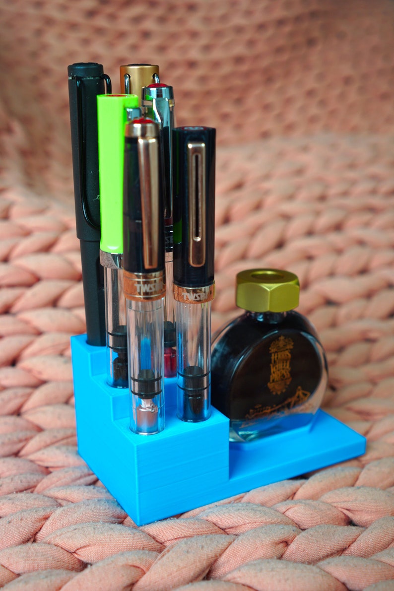 Puede incluir: Un portaplumas de pl&aacute;stico azul con cinco plumas y un frasco de tinta. Las plumas son negras, verdes, plateadas y doradas. El frasco de tinta es transparente con una tapa dorada y dice "Herbs Hill Press" en la etiqueta.