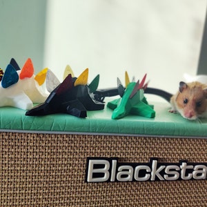 May include: Five colorful guitar picks shaped like dinosaurs are arranged on a green surface. A hamster is sitting on the surface behind the picks. The surface is on top of a brown and beige guitar amplifier with the word "Blackstar" printed on the front.