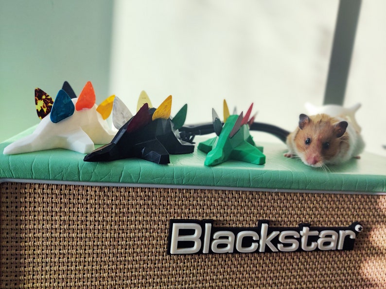 May include: Five colorful guitar picks shaped like dinosaurs are arranged on a green surface. A hamster is sitting on the surface behind the picks. The surface is on top of a brown and beige guitar amplifier with the word "Blackstar" printed on the front.