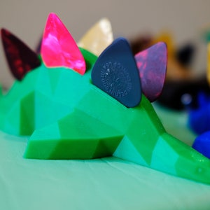 May include: A green, 3D printed dinosaur-shaped guitar pick holder with four guitar picks inserted. The picks are pink, black, red, and beige.