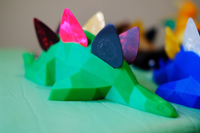 May include: A green, 3D printed dinosaur-shaped guitar pick holder with four guitar picks inserted. The picks are pink, black, red, and beige.