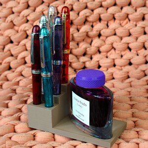 Puede incluir: Un portaplumas gris con cinco plumas estilogr&aacute;ficas de diferentes colores, incluyendo rojo, azul y verde. Una botella de tinta morada con una tapa morada est&aacute; en el portaplumas. El portaplumas est&aacute; sobre una superficie de punto de color melocot&oacute;n.