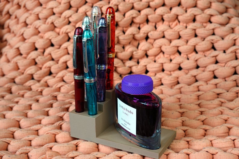 Puede incluir: Un portaplumas gris con cinco plumas estilogr&aacute;ficas de diferentes colores, incluyendo rojo, azul y verde. Una botella de tinta morada con una tapa morada est&aacute; en el portaplumas. El portaplumas est&aacute; sobre una superficie de punto de color melocot&oacute;n.