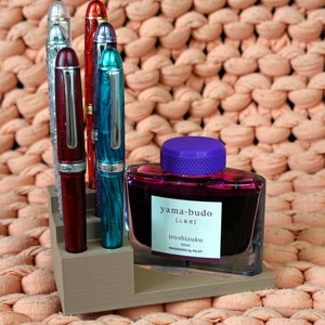 Puede incluir: Un juego de cinco plumas estilogr&aacute;ficas en un soporte de madera, con una botella de tinta morada etiquetada "yama-budo Iroshizuku 50ml PRODUCED BY PILOT".