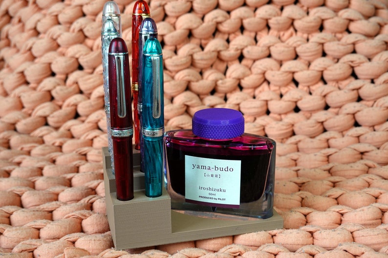 Puede incluir: Un juego de cinco plumas estilogr&aacute;ficas en un soporte de madera, con una botella de tinta morada etiquetada "yama-budo Iroshizuku 50ml PRODUCED BY PILOT".