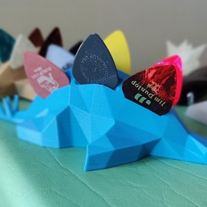 May include: A blue 3D printed guitar pick holder shaped like a dinosaur, with five guitar picks in various colors and designs, including a Jim Dunlop pick with the text "Jim Dunlop" and "Max Grip" printed on it.