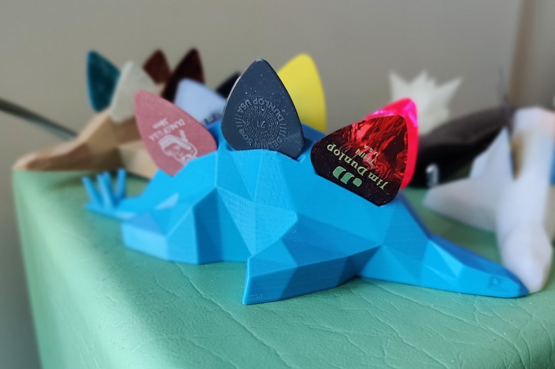 May include: A blue 3D printed guitar pick holder shaped like a dinosaur, with five guitar picks in various colors and designs, including a Jim Dunlop pick with the text "Jim Dunlop" and "Max Grip" printed on it.