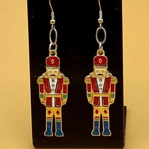 May include: A pair of dangle earrings featuring a festive nutcracker soldier design. The earrings have a red and gold color scheme with blue boots and a white face. The earrings are displayed on a black stand.