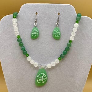 May include: A necklace and earring set featuring green and white beads. The necklace has a green pendant with a white floral design. The earrings are teardrop-shaped, matching the pendant design. The jewelry is displayed on a gray mannequin.