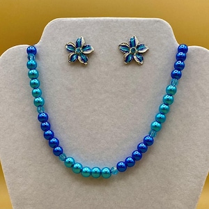 May include: A blue and turquoise ombre pearl necklace with matching flower-shaped earrings. The necklace and earrings are made of iridescent glass beads.