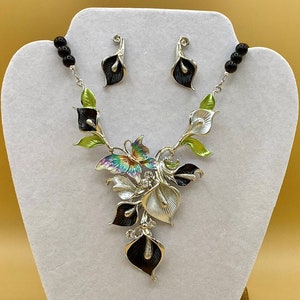 May include: A silver necklace with a black and white flower design and a butterfly pendant. The necklace is accented with green and black beads. The necklace comes with matching earrings.