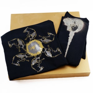 May include: Two black t-shirts with unique designs. One shirt features a circular design of gray animals around a gold eye. The other shirt has a white and gray design. The shirts are presented on a gold box.