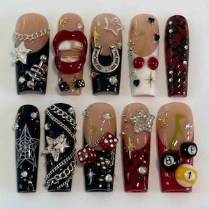 Könnte beinhalten: Ein Set aus zehn langen, künstlichen Nägeln mit verschiedenen Designs. Die Nägel haben schwarze, rote und nudefarbene Basen, verziert mit Charms wie Sternen, Ketten, Würfeln, einem Hufeisen und einem Spinnennetz. Einige Nägel haben ein French-Tip-Design, andere zeigen komplizierte Muster.