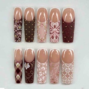 May include: A collection of ten artificial nails with diverse designs. The nails have a natural base with white tips. Designs include hearts, gold studs, floral patterns, and plaid. Colours include brown, pink, and burgundy.