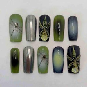 Könnte beinhalten: Ein Set aus zehn künstlichen Nägeln mit verschiedenen Designs. Die Nägel weisen eine Mischung aus Farben auf, darunter metallisches Silber, Grün und Dunkelblau. Einige Nägel haben Libellen- und Blumendesigns, andere haben Farbverläufe.