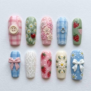 Könnte beinhalten: Zehn künstliche Fingernägel mit verschiedenen Designs, darunter rosa und blaue Karomuster, weiße Spitzenstrukturen, 3D-Blumen, Erdbeeren, Gänseblümchen, ein Marienkäfer, ein kleiner silberner Ring sowie rosa oder weiße Schleifen.