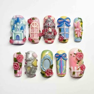Könnte beinhalten: Eine Sammlung von zehn dekorativen Nagelkunst-Designs. Die Designs umfassen ein Schloss, ein Fenster mit Rosen, einen Vogelkäfig, ein Fenster mit einer blauen Schleife, eine rosa Schleife, ein rosa-weiß gestreiftes Design mit Rosen, das Wort "LOVE", einen Bilderrahmen, eine blaue Schleife und ein rosa Design mit Rosen.