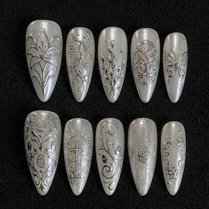 Könnte beinhalten: Ein Set aus zehn langen, mandelförmigen Kunstnägeln. Jeder Nagel ist perlmuttweiß mit aufwendigen silbernen Designs, darunter Blumen, Ranken, Schmetterlinge und Ostermotive. Die Nägel sind auf schwarzem Grund angeordnet.