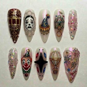 Op de afbeelding: Een set van tien lange, amandelvormige kunstnagels met een circusthema. Ontwerpen omvatten een clowngezicht, een circustent, een heteluchtballon en een verdrietig clowngezicht. Kleuren zijn rood, wit, goud en iriserende glitter.