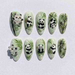 Könnte beinhalten: Ein Set aus zehn mandelförmigen Kunstnägeln. Jeder Nagel zeigt ein Panda-Bär-Design mit Bambusblättern. Einige Nägel haben 3D-Panda-Bär-Akzente. Die Farbpalette umfasst Weiß, Schwarz und Grüntöne.
