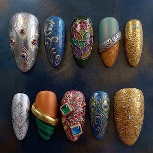 Könnte beinhalten: Eine Sammlung von zehn aufwendig gestalteten Kunstnägeln. Die Nägel weisen eine Vielzahl von Farben auf, darunter Silber, Gold, Blau, Grün und Orange, mit aufwendigen Mustern, Edelsteinen und Texturen. Jeder Nagel zeigt einzigartige Kunstfertigkeit und Design.