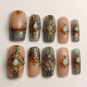 Könnte beinhalten: Ein Set aus zehn künstlichen Fingernägeln mit einer Basis in Nude und Grau. Die Designs umfassen goldene Filigranmuster, geometrische Goldrahmen mit eingebetteten grünen Kristallen und tropfenförmige Opale, die in goldene Akzente gefasst sind.