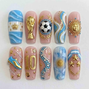 Könnte beinhalten: Ein Set aus zehn künstlichen Fingernägeln mit Argentinien-Motiven. Die Verzierungen umfassen einen goldenen Weltpokal, einen Fußball, die Zahl 10, ein Sonnenemblem, blau-weiße Streifen, goldene Sterne, einen Fußballschuh und blaue Strasssteine.
