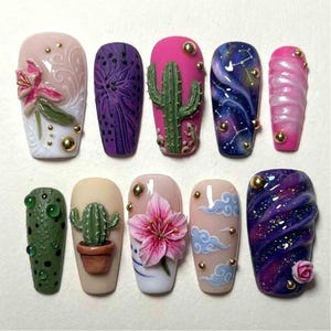 Könnte beinhalten: Eine Sammlung von zehn künstlichen Fingernägeln mit aufwendigem Design. Die Nägel zeigen verschiedene Motive, darunter Kakteen, Galaxien und Blumenmuster. Die Farben umfassen Rosa, Lila, Grün und Blau, mit goldenen Akzenten. Jeder Nagel ist einzigartig und detailliert.