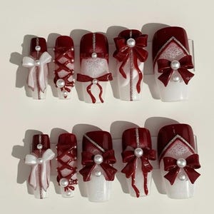 Könnte beinhalten: Ein Set aus zehn künstlichen Nägeln mit weißer Basis und verschiedenen Designs. Einige Nägel haben eine tiefrote Spitze mit Perlenakzenten, während andere weiße Schleifen, rote Kreuzmuster und dekorative Spitze zeigen. Die Nägel sind auf einer weißen Oberfläche angeordnet.