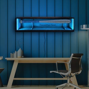Può includere: Una teca in legno per la mazza da baseball con luci LED blu. La teca è montata su una parete blu e ha la scritta "I loved her first" stampata sul davanti.