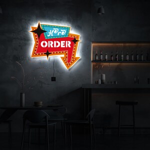 May include: A neon sign with a red arrow pointing to the right. The sign says "Here to Order" in white letters with a black outline. The sign is mounted on a gray wall in a bar setting.