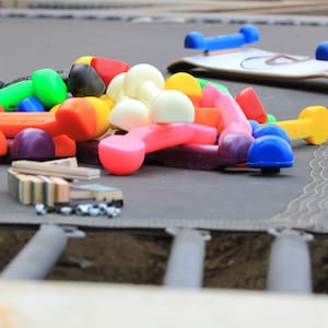 Op de afbeelding: Een verzameling kleurrijke speelgoed dumbbells en andere speelartikelen ligt op een trampoline. De dumbbells zijn in verschillende kleuren, waaronder oranje, geel, groen en blauw. Een speelgoed skateboard met blauwe wielen is ook zichtbaar.