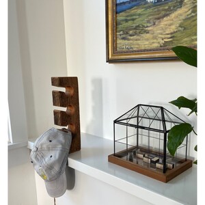 May include: A wooden hat rack with three slots, a gray baseball cap hanging from the rack, a small glass greenhouse with a wooden base, and a framed painting of a landscape with a white building in the distance.