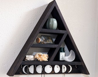 May include: A black triangular shelf with multiple shelves and hooks. The shelf has a moon and crescent moon design. There are several necklaces hanging from the hooks. There is a small green pot on the top shelf, a small glass bottle on the middle shelf, and a larger glass bottle and a white crystal on the bottom shelf.
