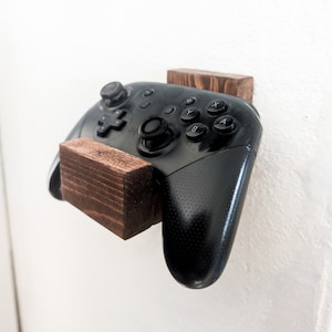 May include: A black video game controller mounted on a wooden wall mount. The controller has a textured black surface and a variety of buttons.