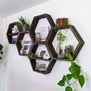 May include: A set of six hexagonal wooden shelves in a dark brown finish, mounted on a white wall. The shelves are arranged in a honeycomb pattern and are filled with various decorative items, including plants, books, crystals, and candles.