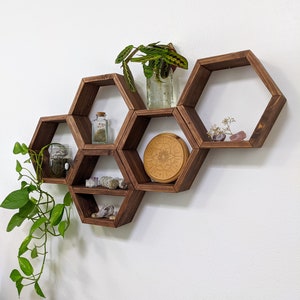 May include: A set of six hexagonal wooden shelves in a dark brown finish, mounted on a white wall. The shelves are arranged in a staggered pattern, creating a honeycomb effect. The shelves are decorated with various items, including plants, crystals, and a wooden mandala.