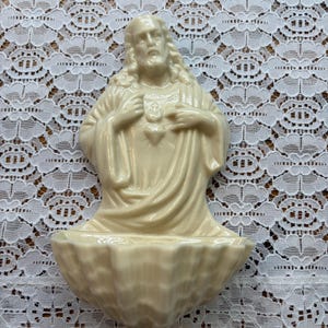 May include: A cream-colored ceramic holy water font featuring a sculpted figure of Jesus Christ with long hair and robes. The figure is positioned above a basin shaped like a scalloped seashell, designed for wall mounting with a glossy finish.