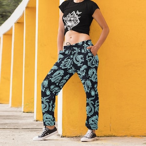 May include: A black t-shirt with a white graphic and dragon print joggers. The joggers are black with a teal dragon pattern. The model wears a grey beanie and black trainers. The logo "Dire Delights" is in the top left corner.