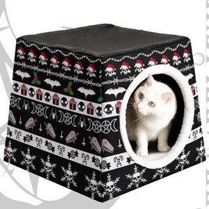 May include: Black and white cat bed with a gothic holiday pattern featuring skulls, bats, and snowflakes. A white cat is inside the bed's opening. The bed has a black mesh top and a white, fluffy interior.